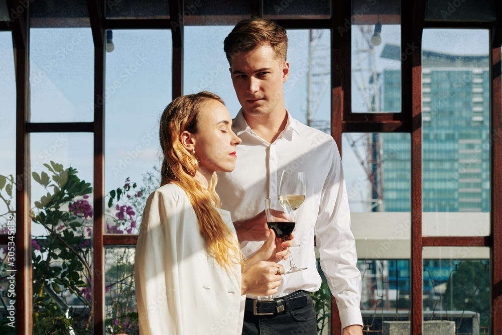 Beautiful young couple with wine glasses standing on restaurant terrace in rays of sun
