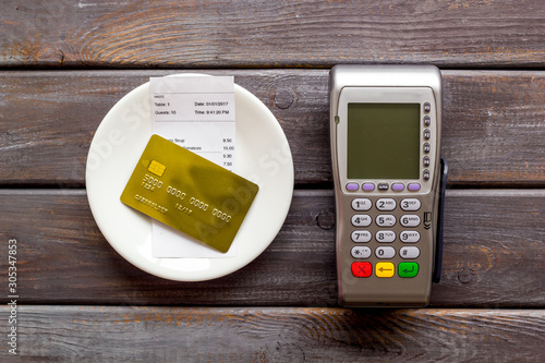 Electronic payments in restaurant. Bank card near aquiring terminal and bill on dark wooden background top view