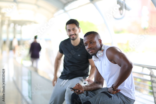 fitness, sport, exercising and lifestyle concept - Two man mix-race in sportswear sitting on relax and stretching leg on step of stadium