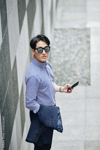 Serious man in sunglasses holding smartphone in one handand jacket in another and looking at camera photo