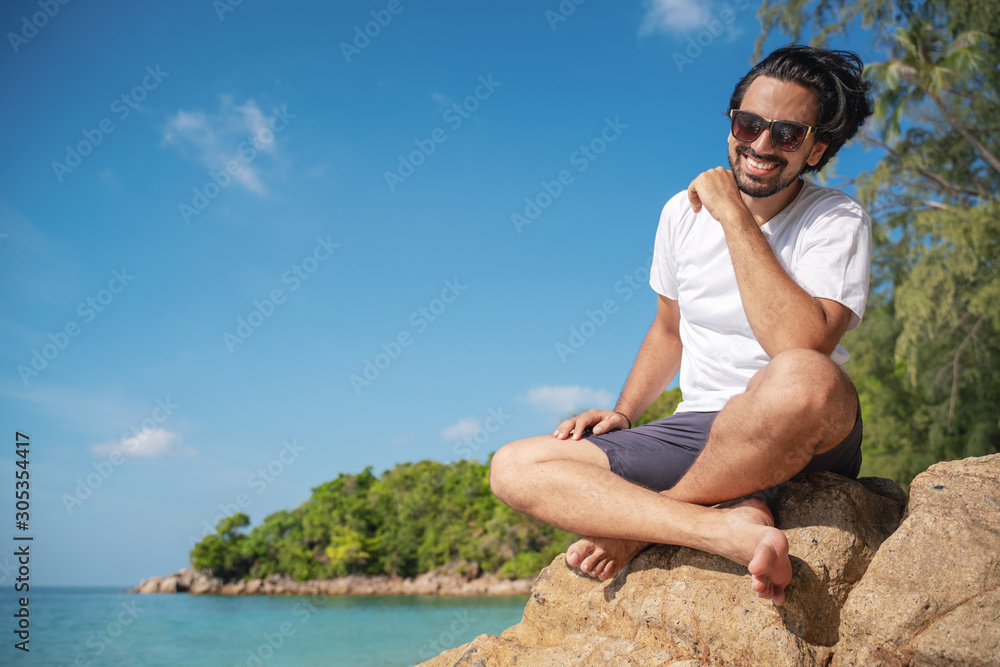 Young fashionable stylish handsome latin man in a white t-shirt and sunglasses on a bright tropical beach, vacation and travel to tropical countries