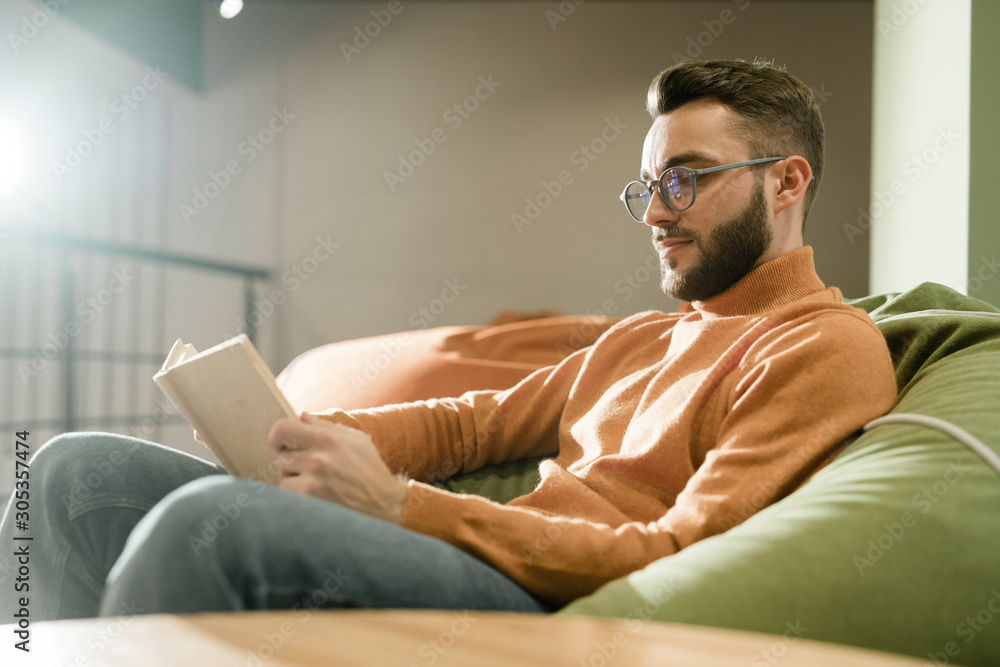 Young relaxed man in casualwear and eyeglasses reading book on comfortable sofa - obrazy, fototapety, plakaty 