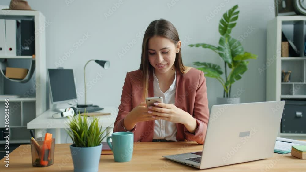 Beautiful lady is using smartphone touching screen smiling sitting at desk in office room alone enjoying gadget. People, workplace and devices concept. Beautiful lady is using smartphone touching screen smiling sitting at desk in office room alone enjoying gadget. People, workplace and devices concept.