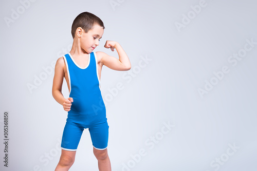 Obraz na plátně Portrait of a little cheerful boy in a blue  wrestling tights shows biceps, looks confidently at him and poses on a white isolated background