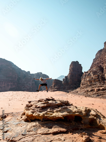 Guy jumping on the edge of a cliff in the desert of Wadi Rum with mountains in the background viewed from the top of the cliff