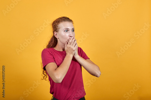 Scared young lady in red shirt covering mouth with hand  looking frightened isolated on orange background in studio. People sincere emotions  sport concept.