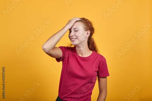 Attractive young lady in red shirt keeping hand on head, smilling, looking happy isolated on orange background in studio. People sincere emotions, sport concept.