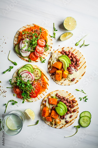 Fotografie Open vegan tortilla wraps with sweet potato, beans, avocado, tomatoes, pumpkin and  sprouts on white background, flat lay, copy space