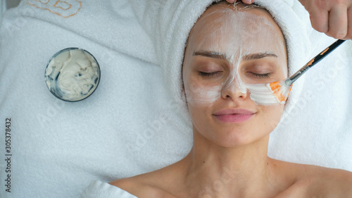 Close up of cosmetologist applies facial clay mask on an young beautiful woman face before ultrasonic cleaning procedure for skin pores and deep moisturizing.
