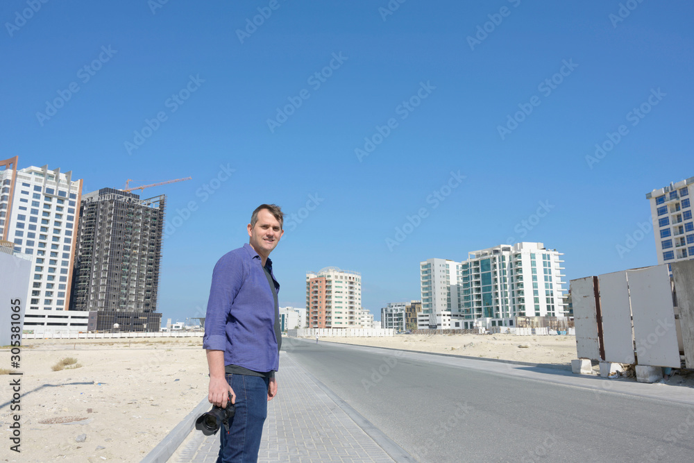 Professional male photographer standing on street holding digital camera smiling, with buildings background over sunny blue sky, Bahrain.