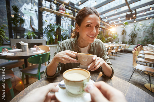 Fisheye view of smiling woman holding coffee cup talking to friend across table in cafe, POV photo
