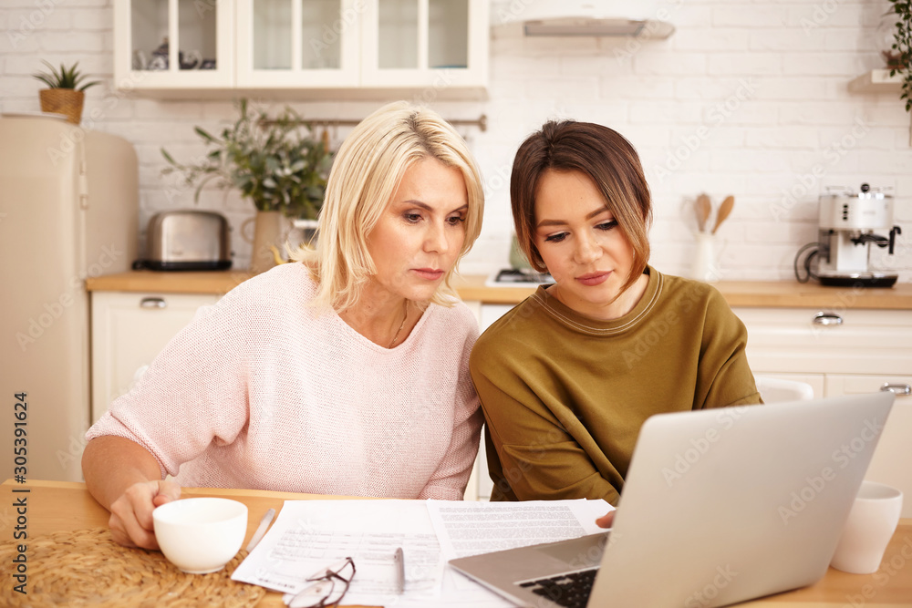 Serious dark haired girl sitting at kitchen table with her middle aged mother using wifi on generic portrable computer, drinking coffee and calculating family budget, planning expensive purchase Serious dark haired girl sitting at kitchen table with her middle aged mother using wifi on generic portrable computer, drinking coffee and calculating family budget, planning expensive purchase