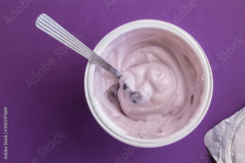 Blueberry yogurt with spoon in plastic cup with foil lid on side isolated on deep purple background - top view