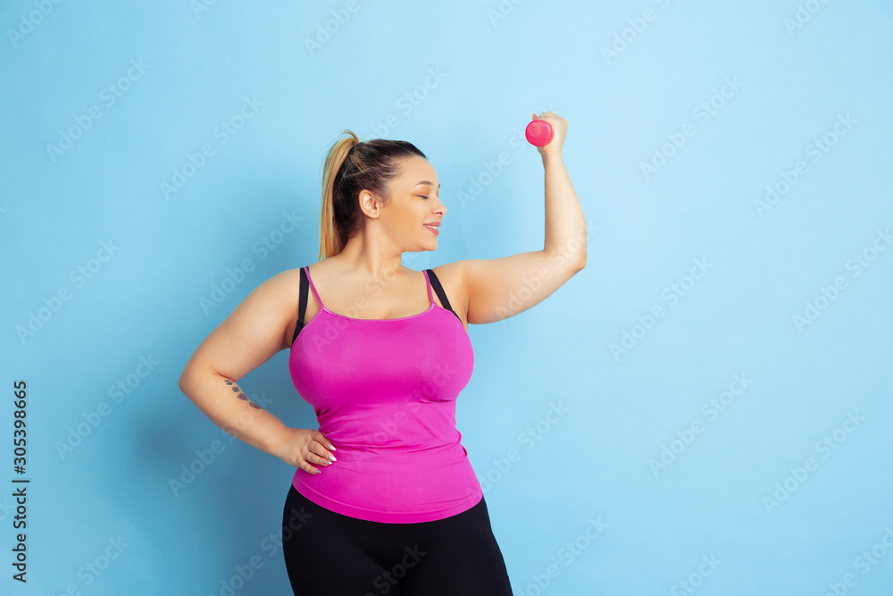 Young caucasian plus size female model's training on blue background. Concept of sport, human emotions, expression, healthy lifestyle, body positive, equality. Training with the weights, copyspace. Young caucasian plus size female model's training on blue background. Concept of sport, human emotions, expression, healthy lifestyle, body positive, equality. Training with the weights, copyspace.