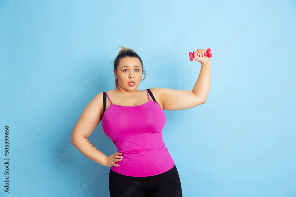 Young caucasian plus size female model's training on blue background. Concept of sport, human emotions, expression, healthy lifestyle, body positive, equality. Training with the weights, copyspace. Young caucasian plus size female model's training on blue background. Concept of sport, human emotions, expression, healthy lifestyle, body positive, equality. Training with the weights, copyspace.