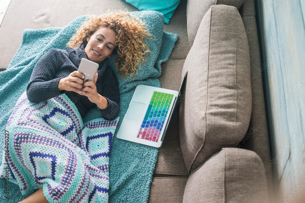 Obraz premium Reaxed caucasian woman type and use mobile phone device lay down on the sofa covered with blue blankets - woman and technology - laptop computer ready for work or onine activity