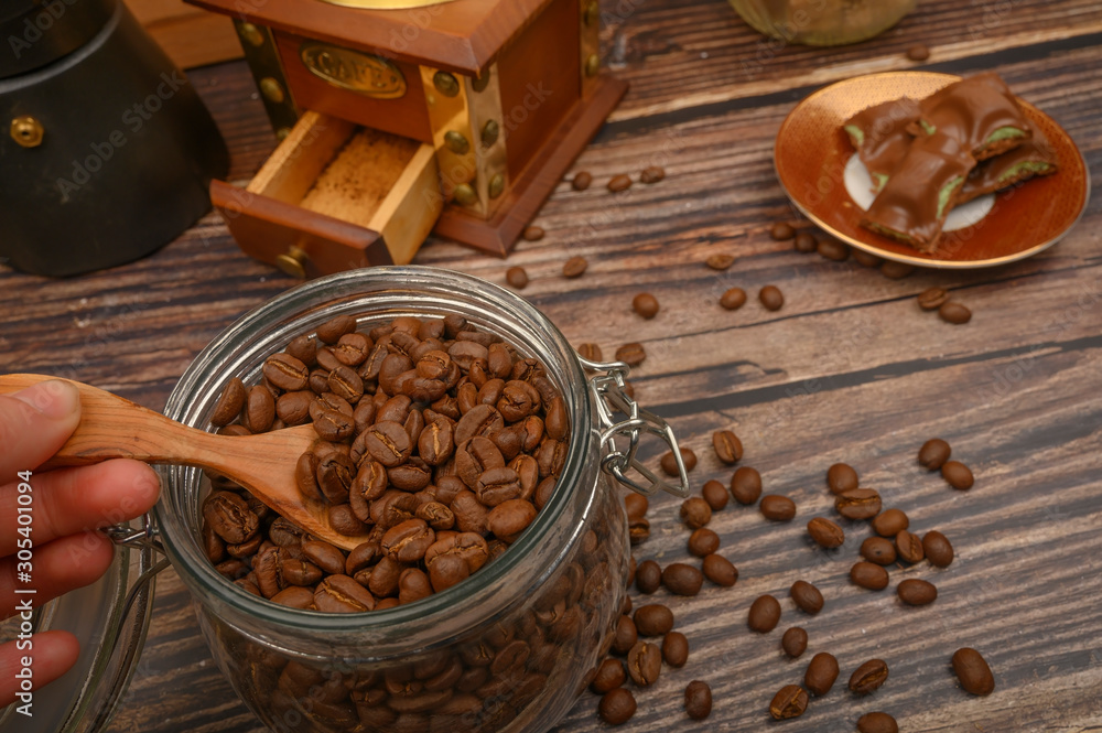 The girl's hand takes a wooden spoon of coffee beans from a glass jar, a coffee grinder, pieces of chocolate on a wooden background. Close up. The girl's hand takes a wooden spoon of coffee beans from a glass jar, a coffee grinder, pieces of chocolate on a wooden background. Close up.