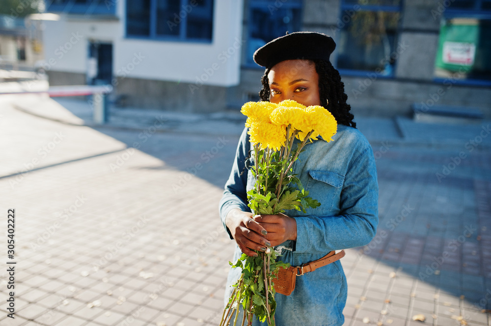 Stylish fashionable african american women in jeans wear and black beret with yellow flowers bouquet posed outdoor in sunny day against blue modern building.