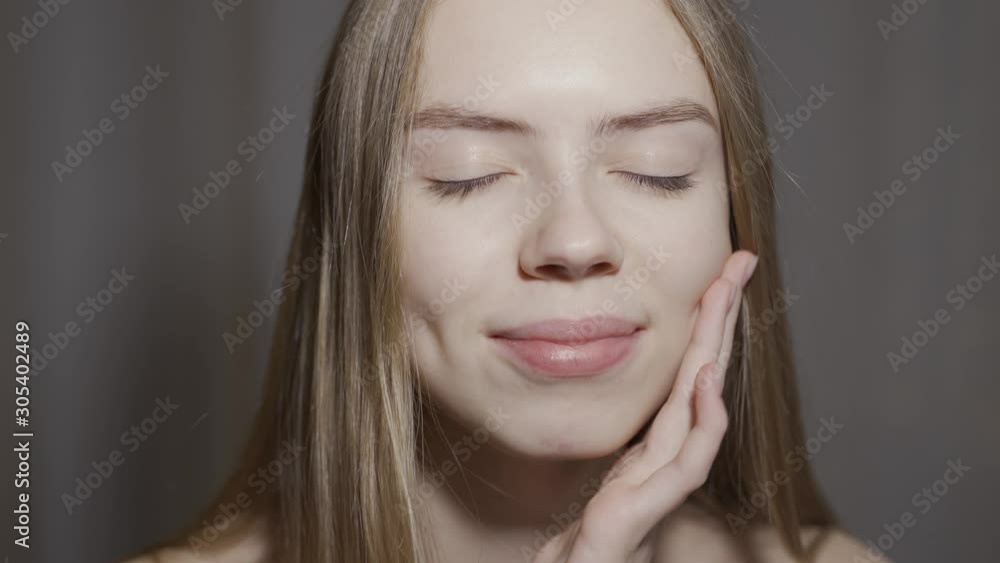 Close-up beauty portrait of young woman with smooth healthy skin, she gently touches her face with her fingers on light grey background and smiles Close-up beauty portrait of young woman with smooth healthy skin, she gently touches her face with her fingers on light grey background and smiles