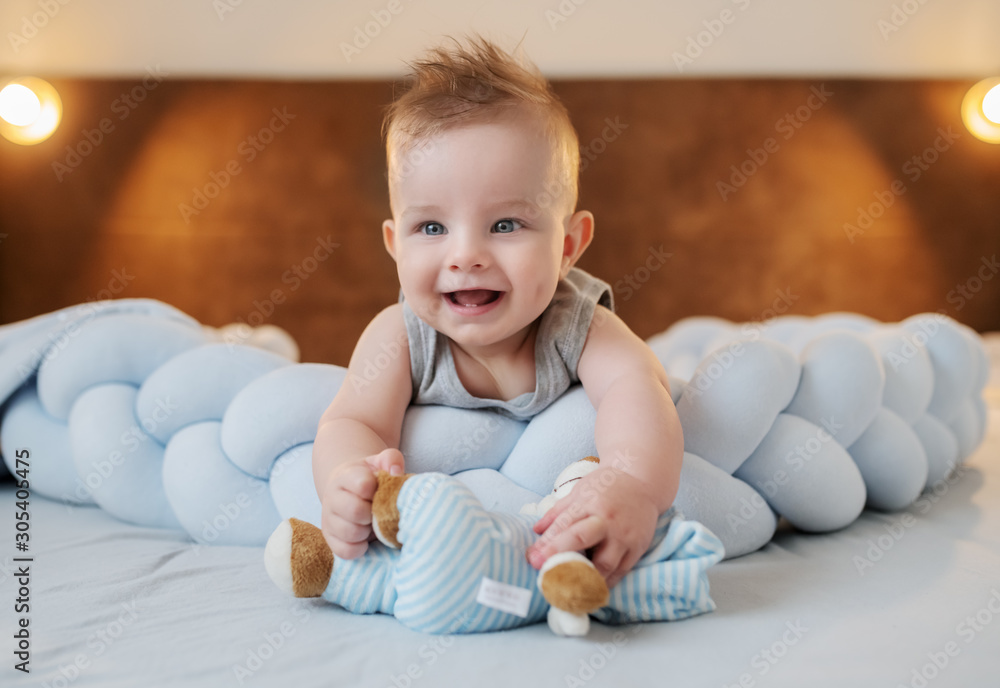 Fototapeta premium Adorable smiling caucasian little baby boy 6 months old lying on stomach on bed in bedroom and holding teddy bear.