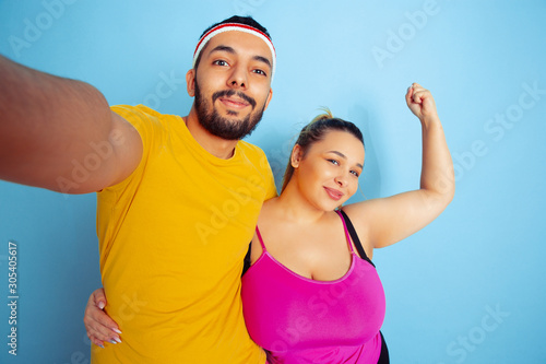 Young pretty caucasian couple in bright clothes training on blue background Concept of sport, human emotions, expression, healthy lifestyle, relation, family. Making selfie together and hugging.