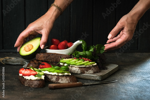 Toast or sandwich with avocado, cheese, strawberries, herbs and seeds on a dark background. Female hands serve a dish. An idea for bruschetta or for a healthy snack. Healthy vegan breakfast.