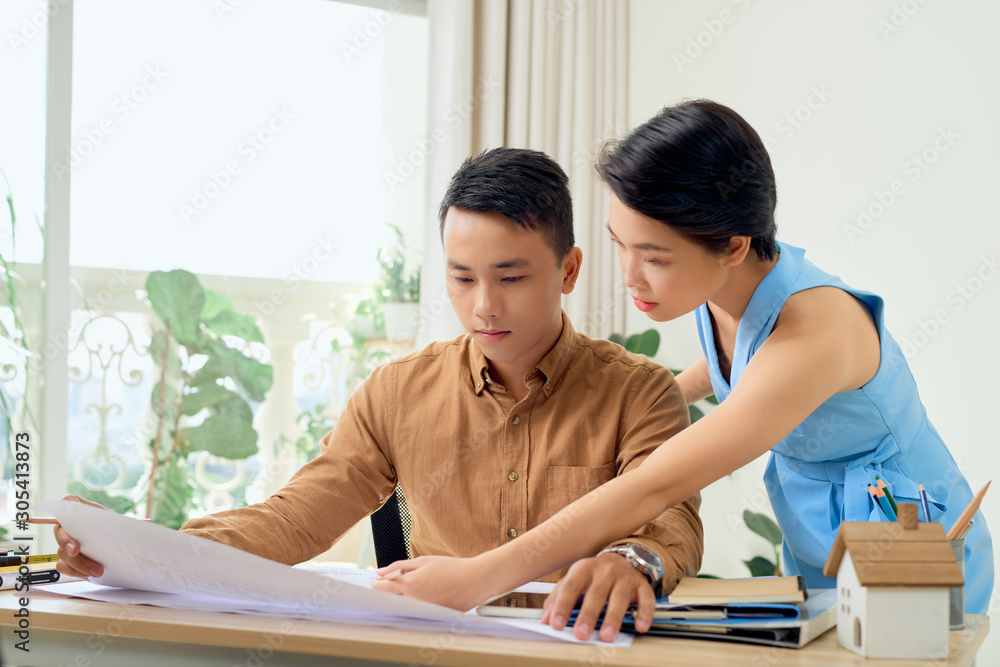 Finding solution together. Confident young man and woman looking at blueprint together while both standing in office Finding solution together. Confident young man and woman looking at blueprint together while both standing in office