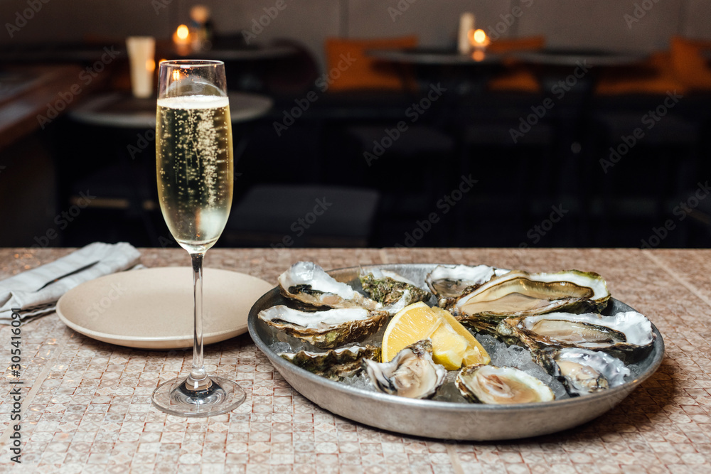 Oysters in a big round metal plate with ice, lemon, and a glass of white dry wine on a stone table and tables on the background. Seafood restaurant
