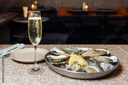 Oysters in a big round metal plate with ice, lemon, and a glass of white dry wine on a stone table and tables on the background. Seafood restaurant