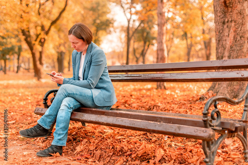 Woman typing text on mobile phone on autumn park bench
