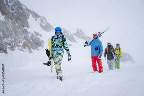 Photo of four sports people with skis and snowboard walking in winter resort