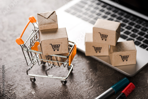 selective focus of toy shopping cart with small carton boxes near laptop, e-commerce concept photo