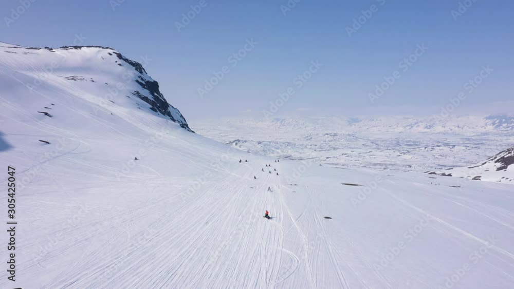 Aerial view of snowy mountain landscape. Sun and blue sky. Following trail towards group of snowmobiles. MOVING FORWARD. Aerial view of snowy mountain landscape. Sun and blue sky. Following trail towards group of snowmobiles. MOVING FORWARD.