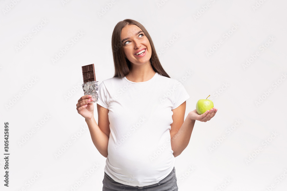 Pregnant Girl Choosing Between Chocolate And Apple Standing, Gray Background Pregnant Girl Choosing Between Chocolate And Apple Standing, Gray Background