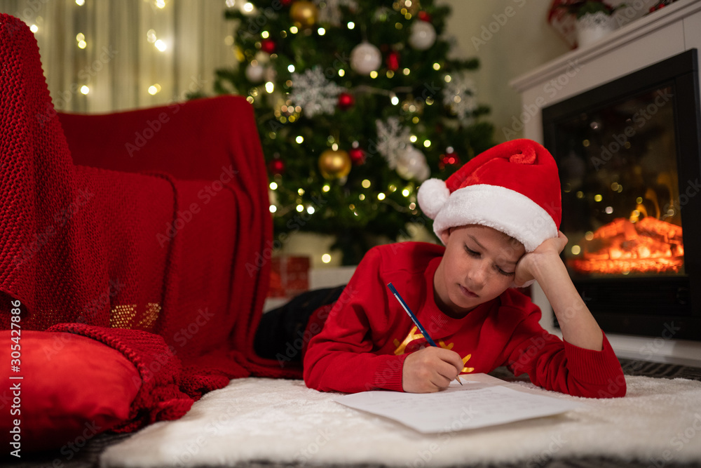 the boy lies under the Christmas tree near the fireplace and writes a letter to Santa Claus the boy lies under the Christmas tree near the fireplace and writes a letter to Santa Claus