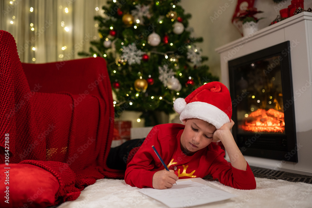 the boy lies under the Christmas tree near the fireplace and writes a letter to Santa Claus the boy lies under the Christmas tree near the fireplace and writes a letter to Santa Claus