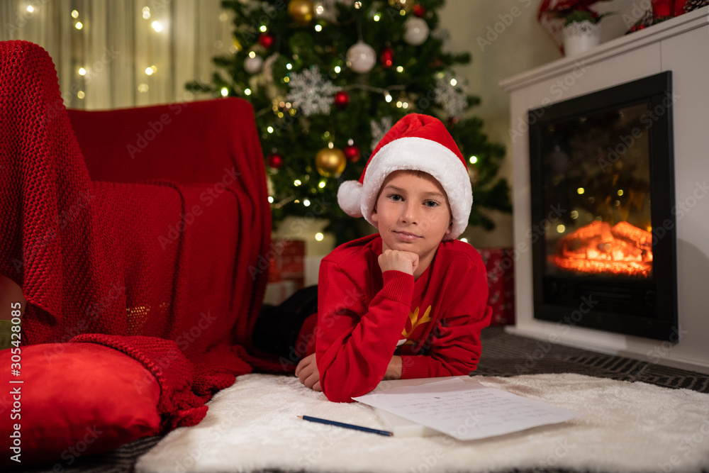 the boy lies under the Christmas tree near the fireplace and writes a letter to Santa Claus the boy lies under the Christmas tree near the fireplace and writes a letter to Santa Claus