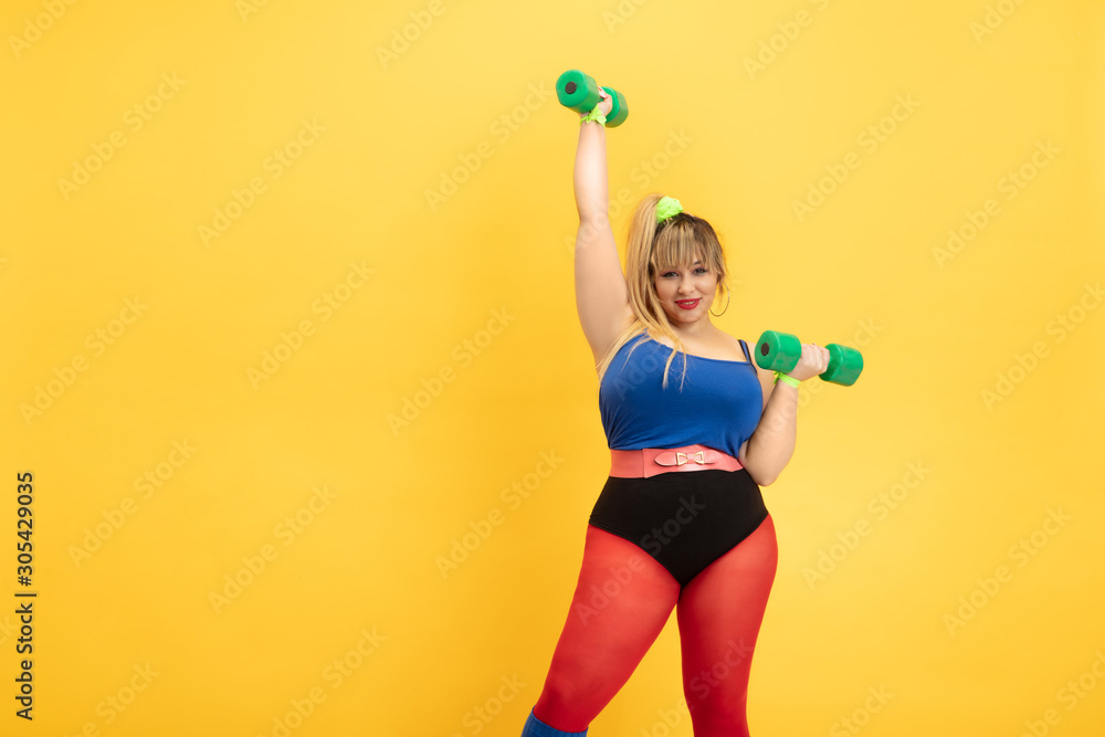 Young caucasian plus size female model's training on yellow background. Stylish woman in bright clothes. Copyspace. Concept of sport, healthy lifestyle, body positive, fashion. Practicing with weights Young caucasian plus size female model's training on yellow background. Stylish woman in bright clothes. Copyspace. Concept of sport, healthy lifestyle, body positive, fashion. Practicing with weights