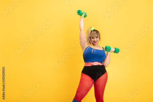 Young caucasian plus size female model's training on yellow background. Stylish woman in bright clothes. Copyspace. Concept of sport, healthy lifestyle, body positive, fashion. Practicing with weights