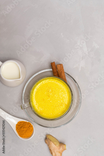 Traditional Indian drink Turmeric Golden Milk in a glass cup with ingredients cinnamon sticks, turmeric powder, ginger root and milk on grey concrete table