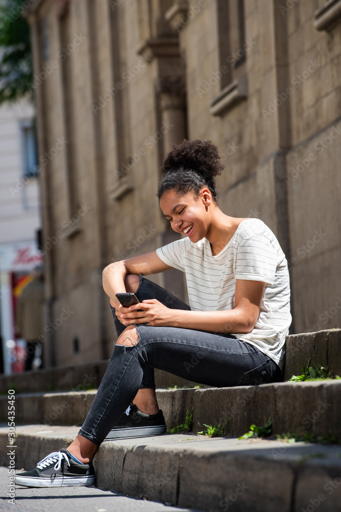 smiling young african american girl sitting outside looking at mobile phone