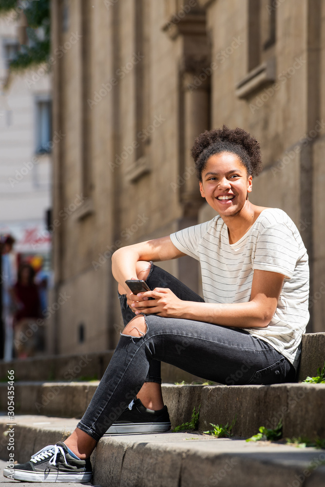 Obraz premium happy young african american girl sitting outside in city with cellphone