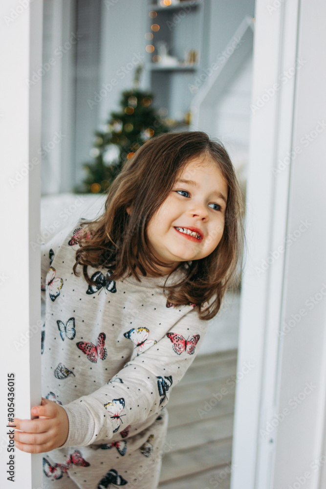 Little happy brunette girl in cozy pajamas looks from children bed room, christmas time Little happy brunette girl in cozy pajamas looks from children bed room, christmas time
