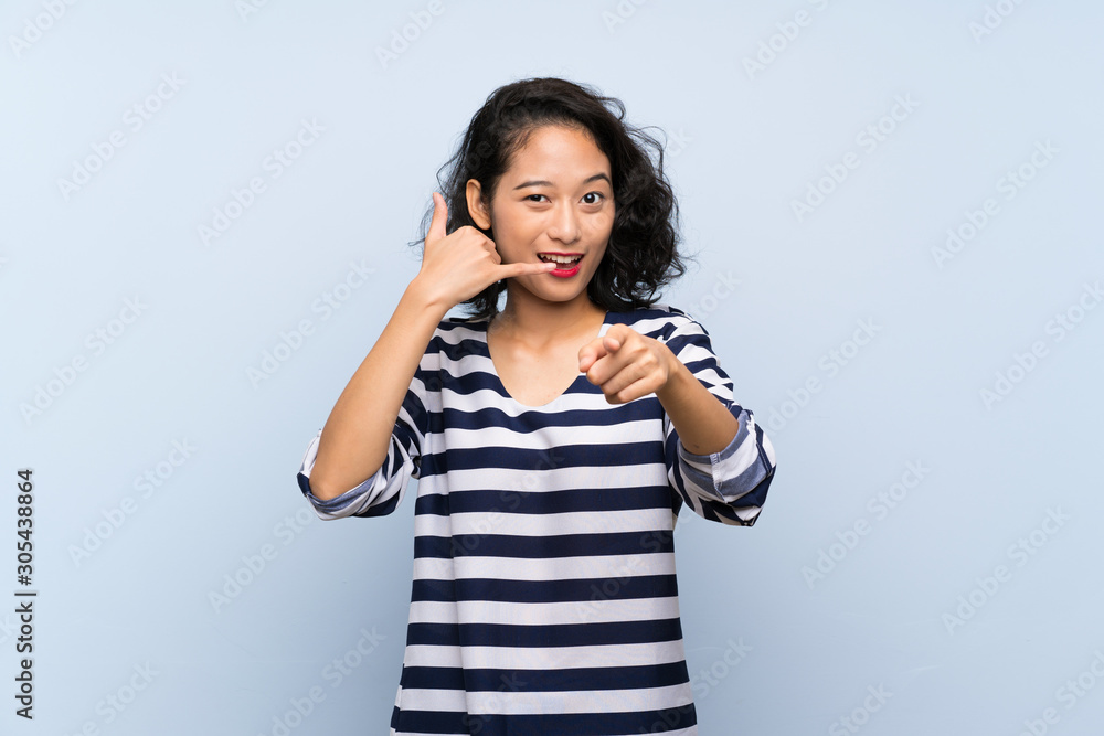 Asian young woman over isolated blue background making phone gesture and pointing front Asian young woman over isolated blue background making phone gesture and pointing front