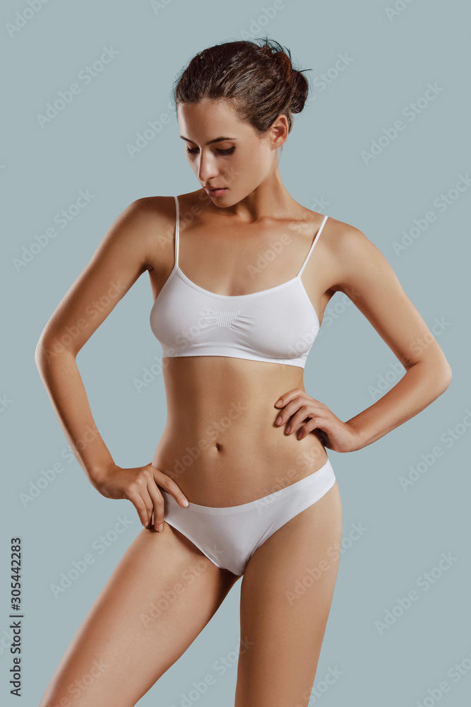Fototapeta premium Young woman in white underwear, with bundled hair, hands on hips, posing against gray background. Plastic surgery, aesthetic cosmetology. Close-up.