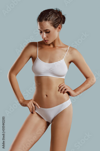 Young woman in white underwear, with bundled hair, hands on hips, posing against gray background. Plastic surgery, aesthetic cosmetology. Close-up.