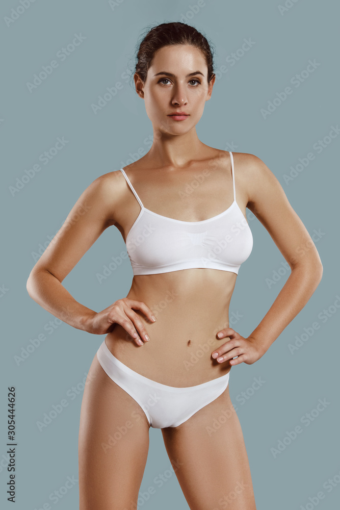 Young woman in white underwear, with bundled hair, hands on hips, posing against gray background. Plastic surgery, aesthetic cosmetology. Close-up. Young woman in white underwear, with bundled hair, hands on hips, posing against gray background. Plastic surgery, aesthetic cosmetology. Close-up.