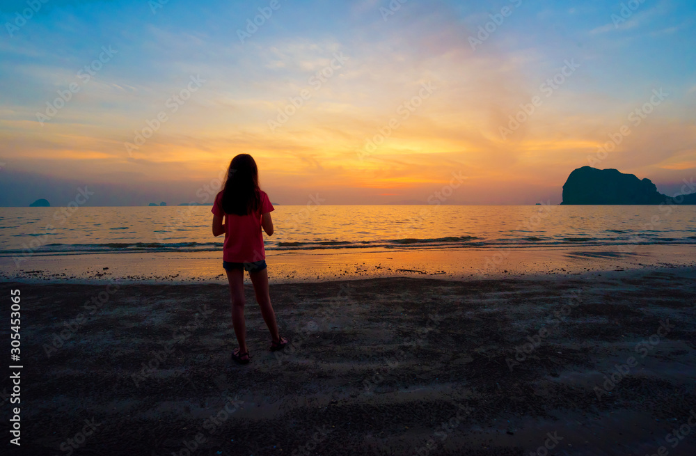 Lonely Asian teenager girl standing on the beach watching sunset sky in the evening. Back of a single sad teen standing alone on the beach near the coast of the sea. Pakmeng beach, Trang, Thailand.