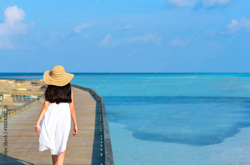 Back of young Asian girl walking on wooden bridge over clear blue sea. Teenager girl wearing sun hat or straw hat and white dress enjoy freedom and fresh air. Outdoor summer vacation travel concept.