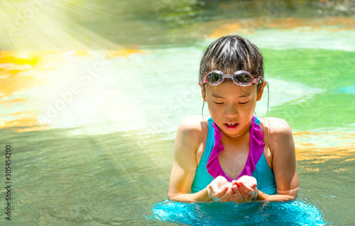 Pretty Asian kid with perfect skin under sun light in swimming pool. Portrait of preteen girl in swimsuit with swimming goggles looking at water in hands in pool side. Summer holiday vacation concept